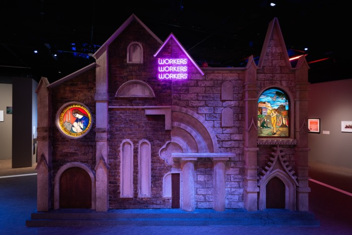 Scaled reproduction of a Medieval church, with stained glass windows and a neon sign which reads ‘workers workers workers’