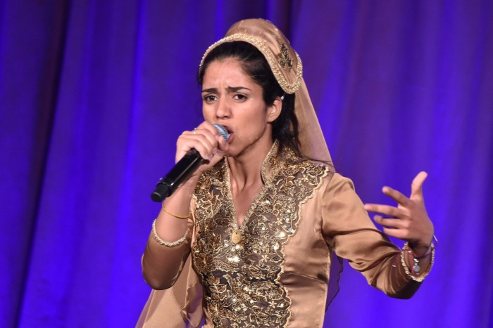 A woman in Afghan clothing and headgear holds up a microphone, gesturing with her free hand