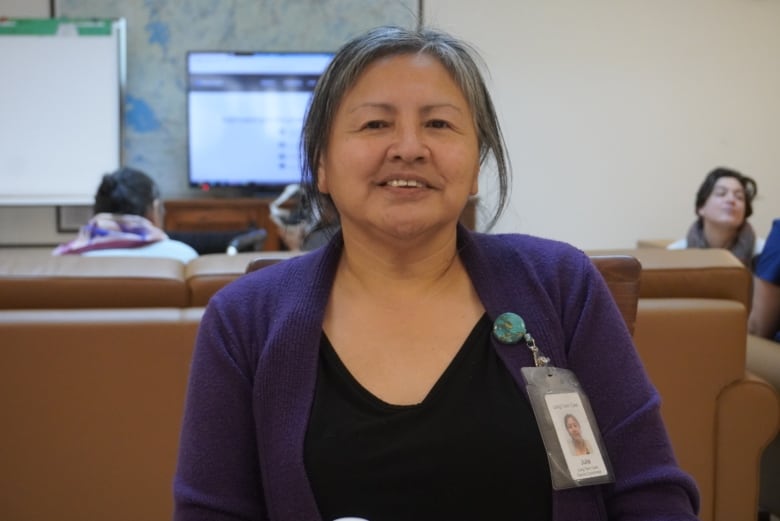 Woman in purple cardigan with staff tag sits, looks at camera.