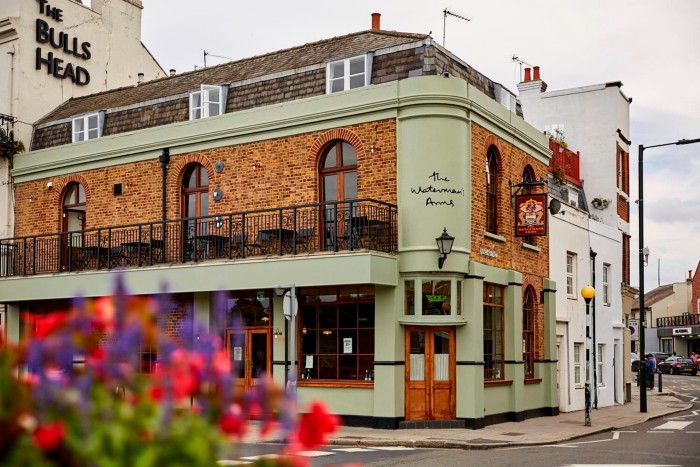 The red-brick and green-painted facade of The Waterman Arms, a pub in a corner building. In the bottom-left foreground are red and purple flowers 