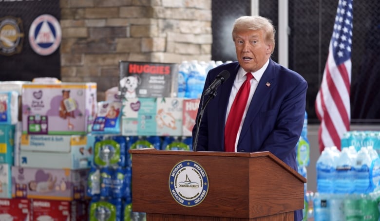 A politician in a blue suit and red tie stands at a podium in front of relief supplies.