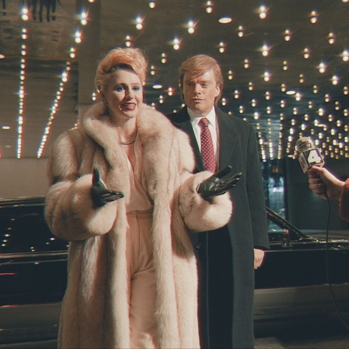 A smiling blonde woman in beige fur coat holds out her glove-clad hands as she speaks into a microphone. Next to her stands a man in black coat over suit and tie. Behind them, lights glint on a limousine