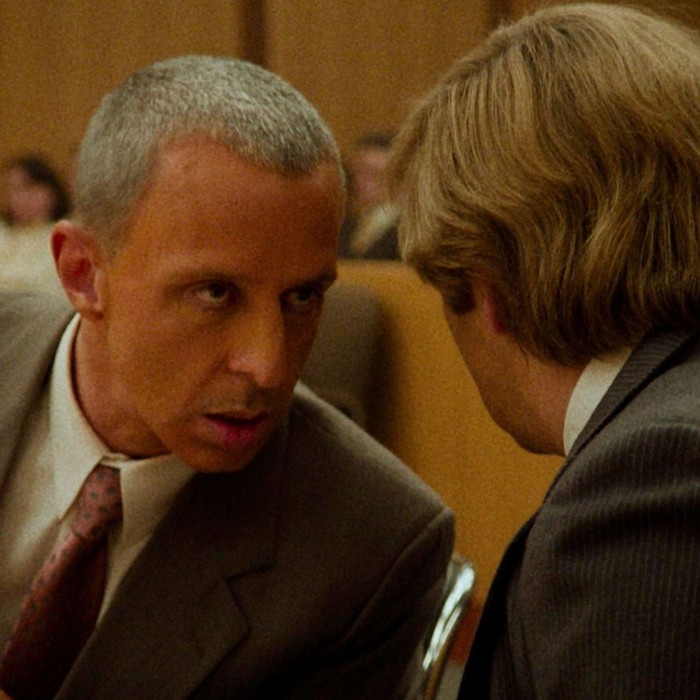 A serious-looking man with cropped grey hair leans in to talk to another man, who has his back to the camera