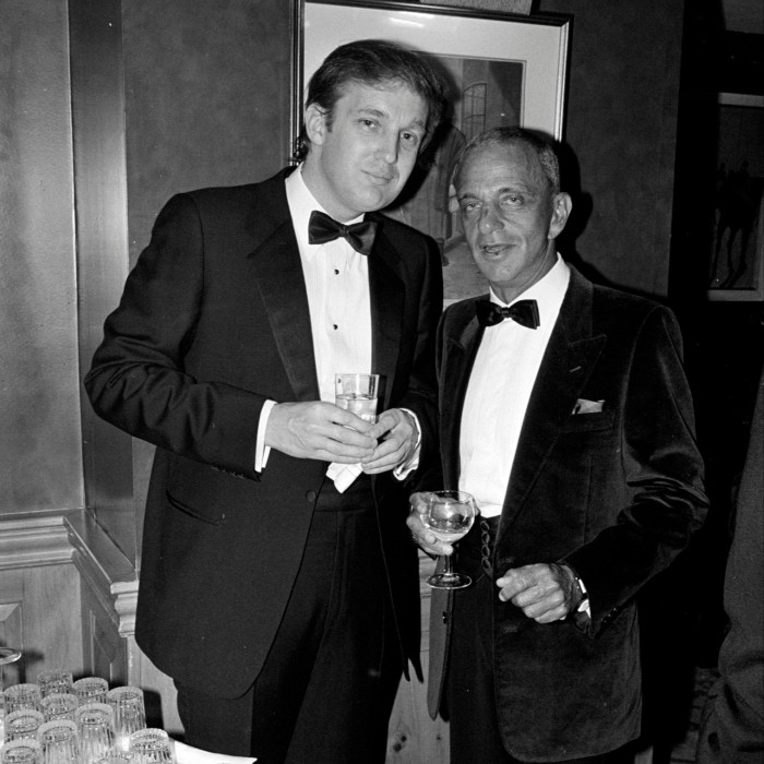 Two men in tuxedos pose for a photo at a party. Both hold a glass. Trump, who is smiling, is the younger and taller of the two