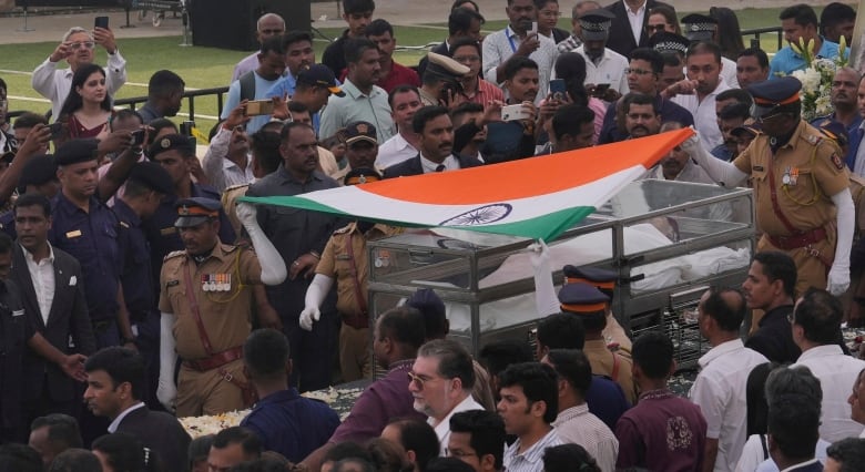 A clear casket draped with a flag is carried through a crowd of people. 