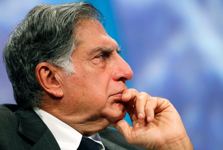 Close-up of an older man's face in profile view, with a blue background. He is wearing a suit and is leaning his chin slightly against his left hand. 