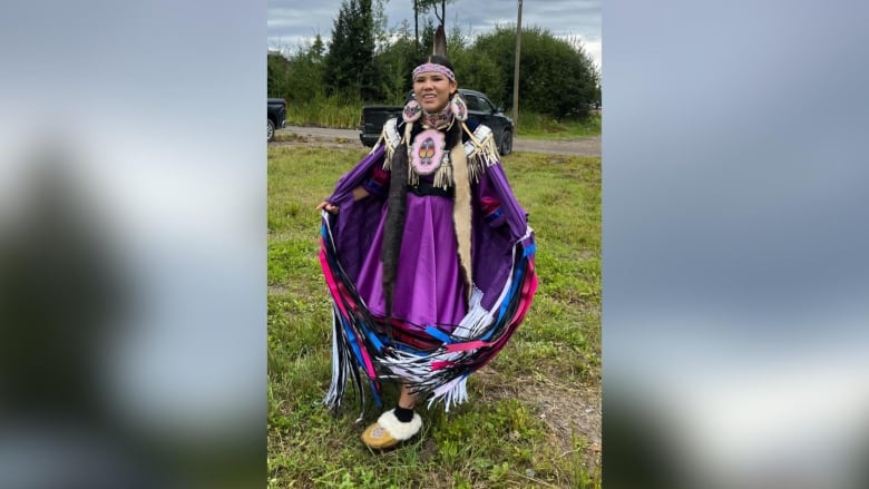 A person stands outside wearing traditional powwow regalia. They are smiling.