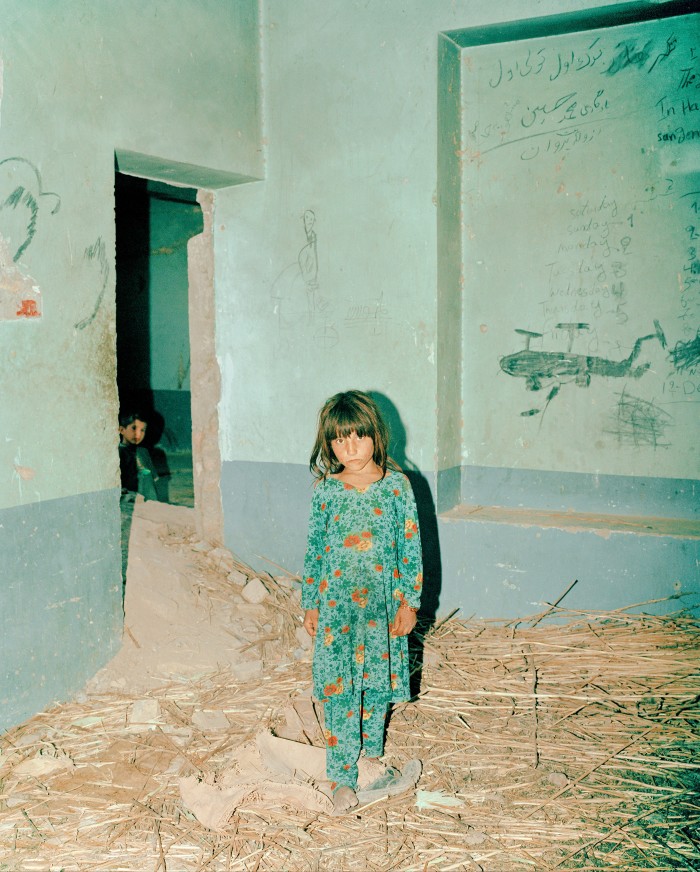 A young girl in a green floral dress stands alone in a bare, dusty room with straw scattered on the ground