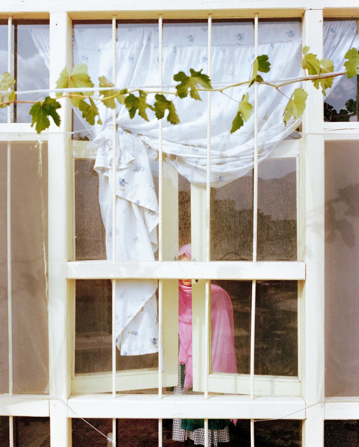 A girl partially obscured by a window stands behind white bars and floral curtains inside a room