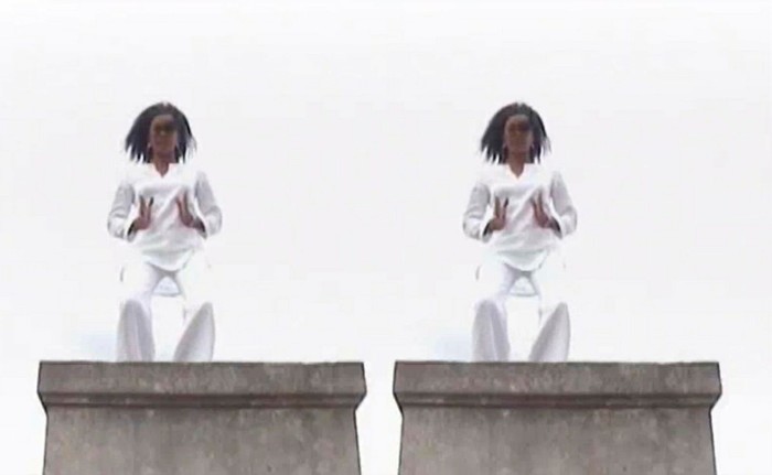 A woman in a loose-fitting white long-sleeved T-shirt and matching white loose-fitting trousers dances on a platform. Her image is repeated so it looks like there are two identical women dancing together 