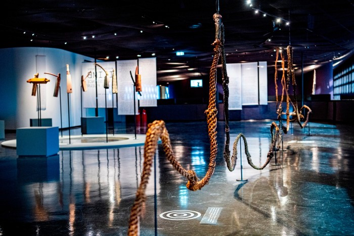 Photograph of an art exhibition containing a number of sculptural objects, including what looks like a long, thick rope made of wickerwork, snaking through the gallery on stands, and various three-dimensional sculptures on plinths 