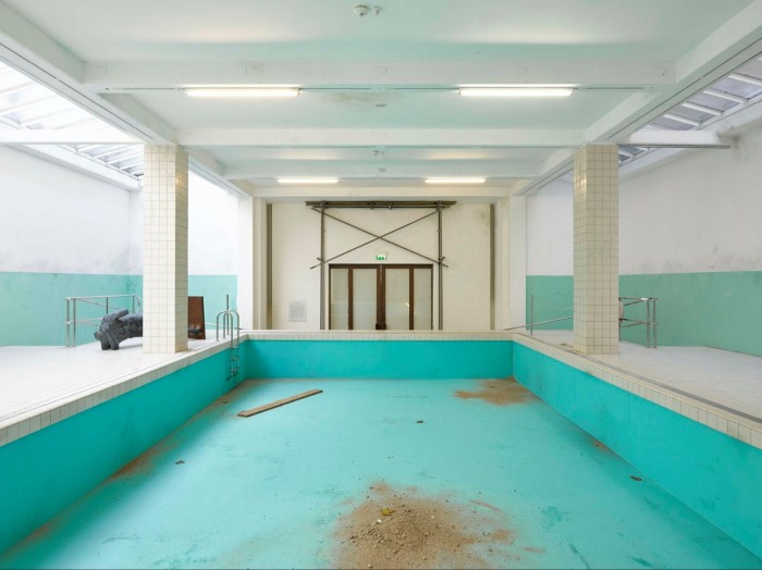 Recreation of an empty swimming pool in an art gallery, with the base coloured turquoise but with pieces of sand, mud and wood scattered around the floor of the pool