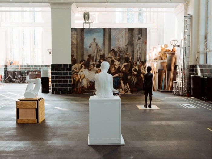 Photograph of an artists studio, with various white and black sculptures in front of a large reproduction of a neo-Renaissance painting depicting decadent Romans