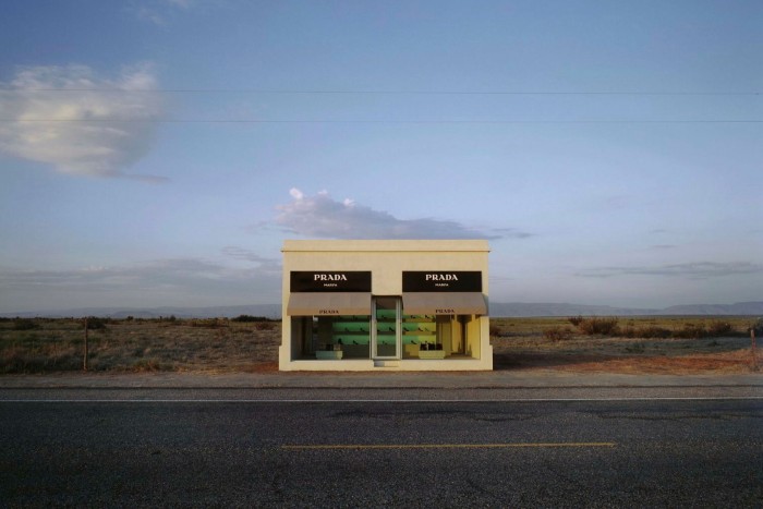 A lone, one-storey building on a roadside in a deserted American landscape, with a shopfront sign that reads ‘Prada Marfa’. In the window you can see shelves containing luxury shoes and handbags