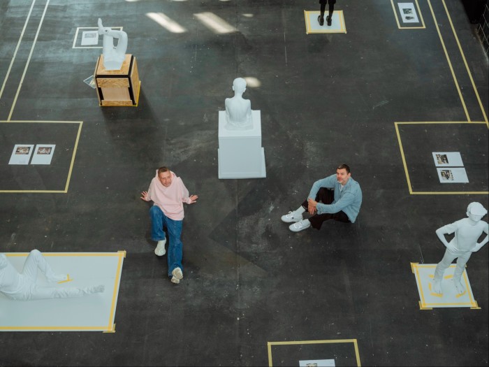 Two men sitting on the floor of an artists studio, surrounded by white plaster sculptures, some of which are on plinths