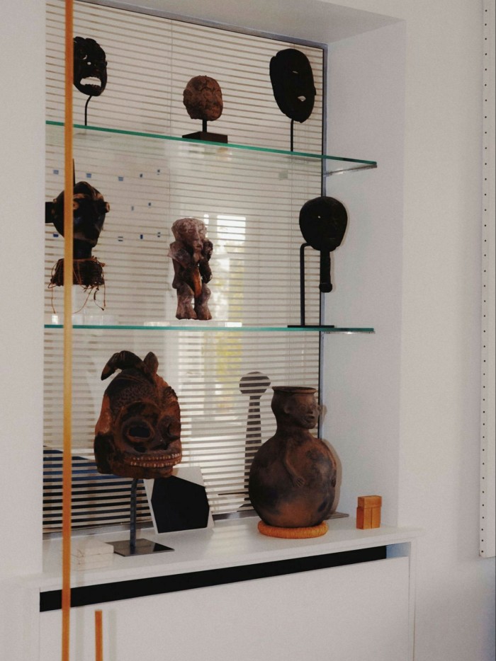 Glass shelves against a mirrored alcove with white walls, displaying what looks like old African art sculptures