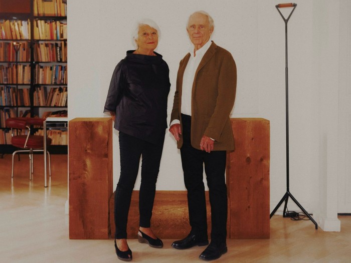 An older couple, both with white hair, in front of a wooden-looking sculptural work comprising three rectangular structures