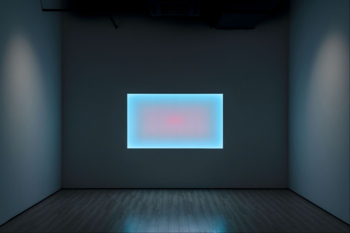 Photograph of a darkened, white-walled art gallery, with a rectangle lit on the wall by a projector. The lit rectangle is coloured pink in the middle and light blue on the edges 