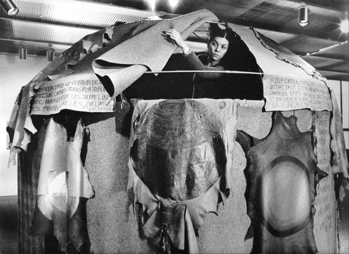 Black and white early 1970s photo of a woman poking her head out of the top of a dome-shaped tent, made from leather and felt, with various French words scribbled on the flaps of the tent