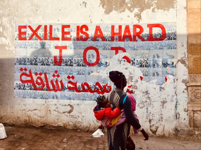 Poster on a wall saying ‘Exile Is A Hard Job’ in red font, translated into Arabic beneath. An African woman with a baby strapped on her back walks past the poster