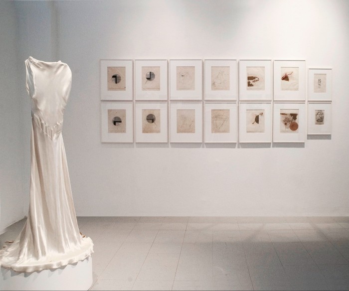 A long, white dress on a headless mannequin on a stand in a white-walled art gallery, with 14 framed pictures in the background, arranged in two horizontal rows