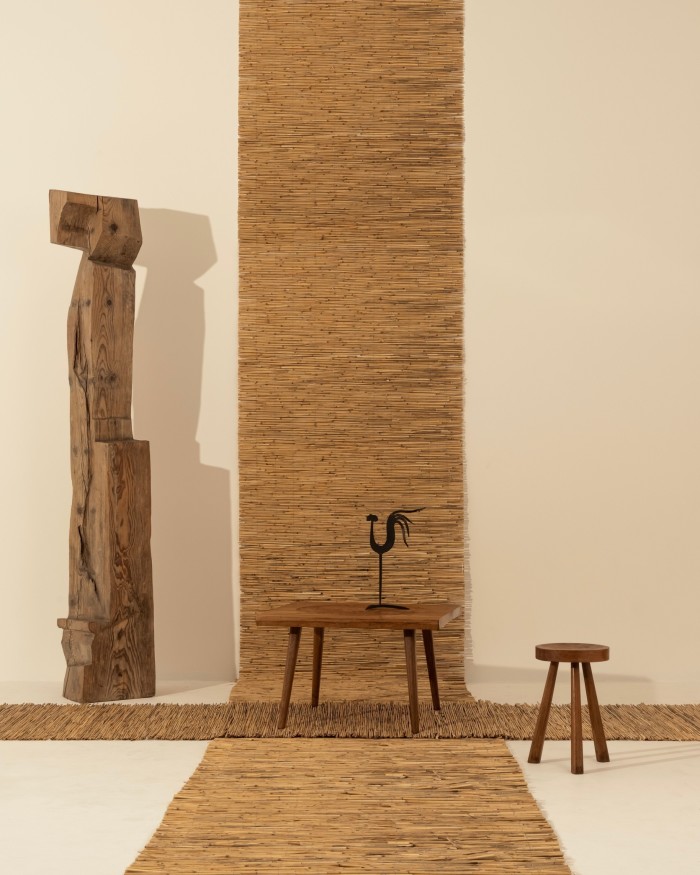 Wooden table and wooden stool, on top of what looks like two rattan carpets, with a wooden abstract sculpture to the side. On top of the table is a metal candle holder