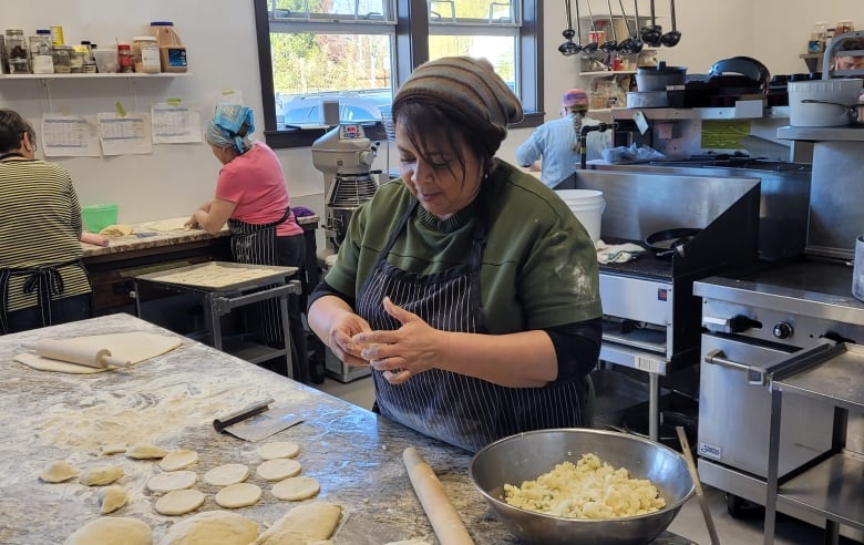 A woman in an apron is seen working with small portions of dough in a commercial kitchen, with other people working in the kitchen behind her.