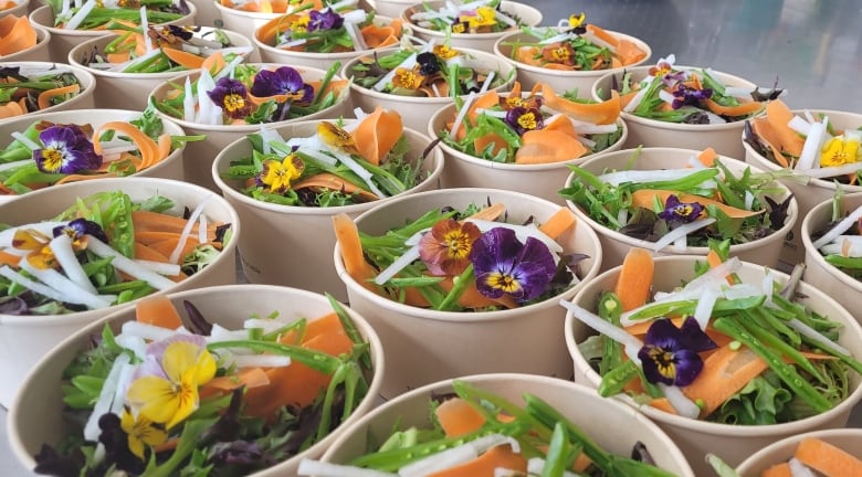 A close-up image showing containers of a bright, colourful salad of greens, shaved carrots and edible flowers is seen laid out a commercial kitchen.