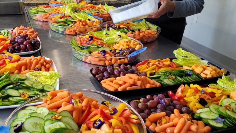 Brightly coloured platters of cut-up carrots, cucumbers, bell pepper, lettuce, melon and grapes, garnished with edible flowers are assembled in a commercial kitchen space.