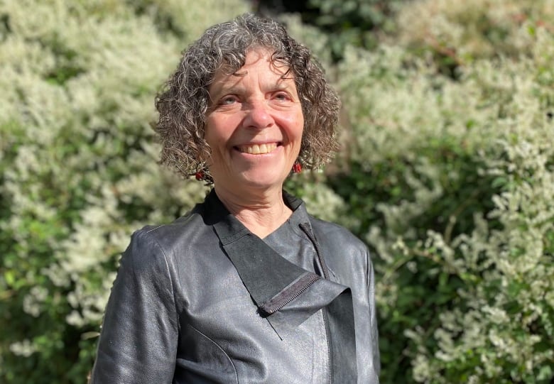 An older woman stands smiling outdoors, next to a flowering bush.