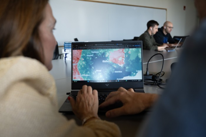 A close-up view of a woman working on a laptop displaying a detailed geographical map, with two colleagues in the background seated at a conference table