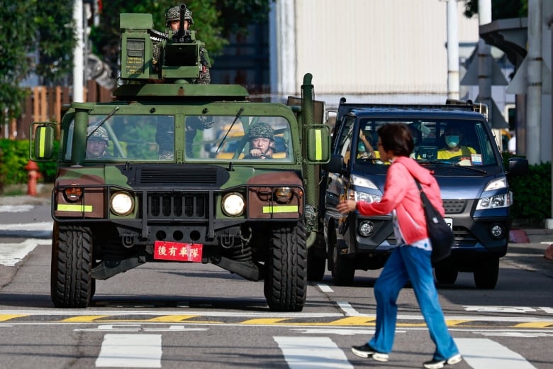 A civilian woman walks in a cross walk in front of a stopped military vehicle topped by a manned gut turret.