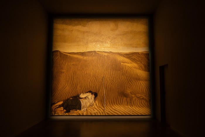 A painting shows a man and a woman lying together and embracing at the foot of a large sand dune; a wooden stick rests against the painting