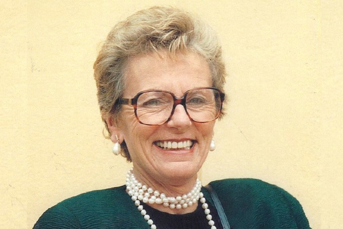 A women with short blonde hair wears large, dark-framed glasses, pearl earrings and a pearl necklace. She is smiling broadly 