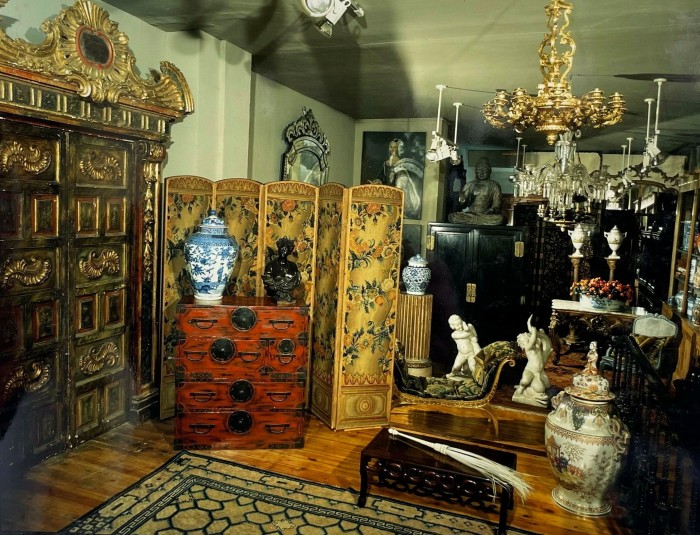 A dark room is filled with furniture and accessories, including gold and crystal chandeliers, ornate ceramic vessels, marble figurines, a floral folding screen and a large and intricate, gold-toned cabinet 