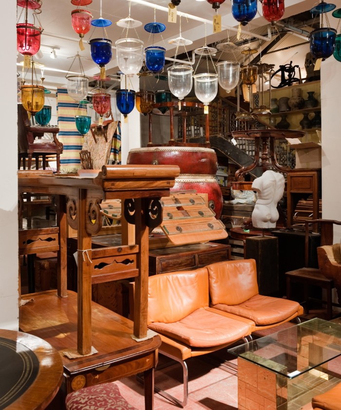 A storeroom is stacked floor-to ceiling with pieces of furniture, some stacked on top of one another, including numerous chairs and tables. The ceiling is hung with a number of lanterns, in clear, blue and red glass 