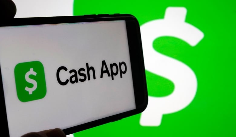 The green and white Cash app logo is seen on a phone being held up at an angle in front of a bigger Cash App logo, which is a green square with a white dollar sign. 