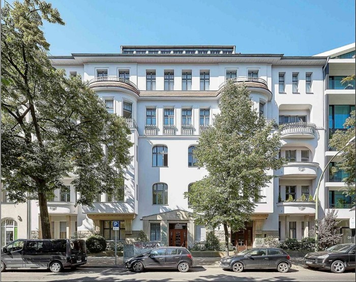 The facade of a multi-story building painted a light, clean white. A few trees and parked cars line the street in front