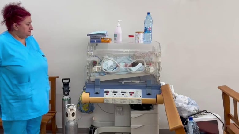 A woman in blue scrubs stands next tp a baby in an incubator