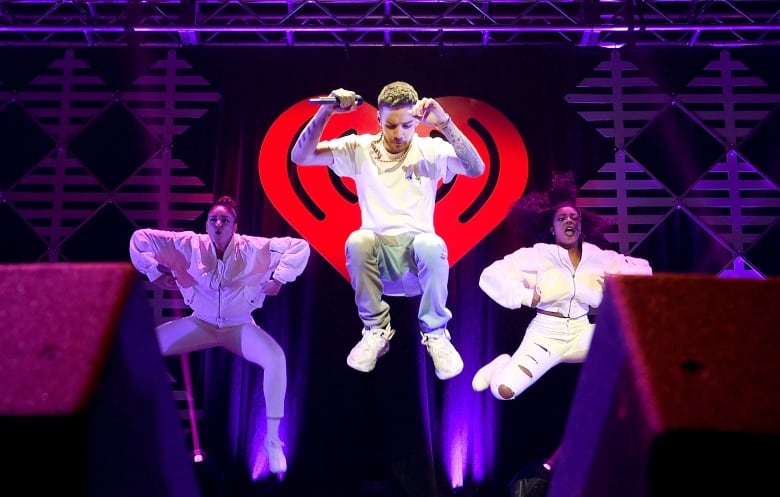 A bearded young man wearing white and holding a microphone jumps onstage while two women behind him perform dance moves.