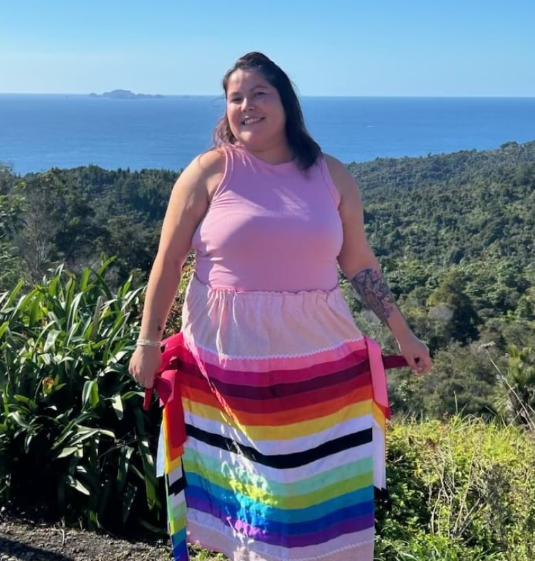 A woman poses in a pink sleeveless shirt and colourful ribbon skirt, with a treed landscape and blue ocean in the background.  