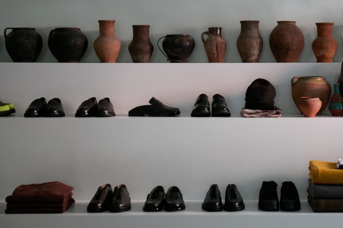 An array of smart shoes on a series of white shelves, on a white wall, which are presented alongside classic-looking pieces of pottery and earthenware jugs