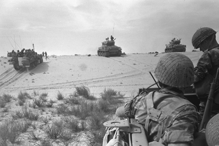 Two soldiers in battle fatigues and helmets watch as troops advance up a sandy incline