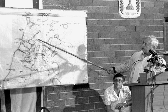 A silver-haired man stands at a lectern, pointing to a map with a stick