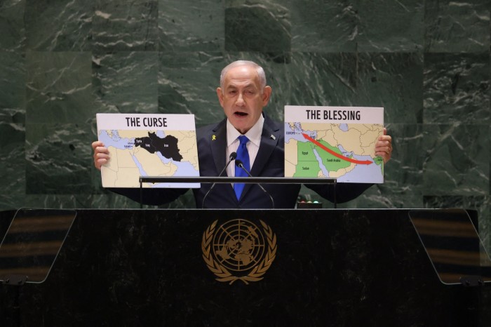 A grey-haired man in dark suit and tie stands at a podium, holding up two small placards, both with maps. One says ‘The Curse’ and the other says ‘The Blessing’