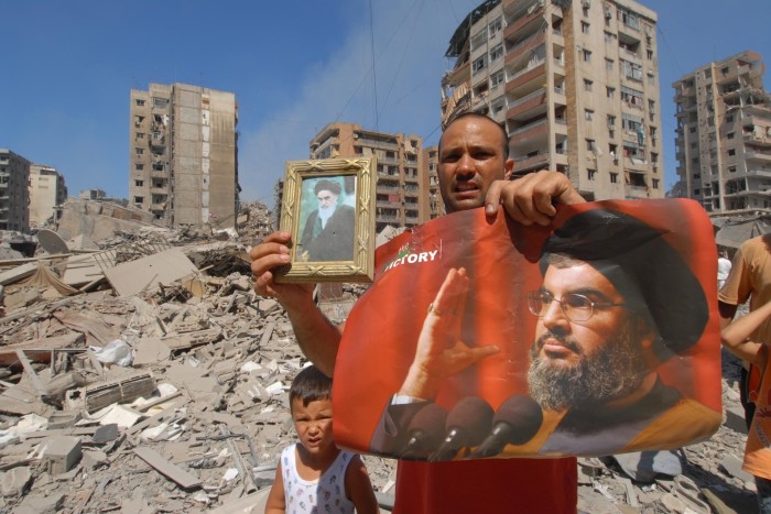 A man stands next to a small child. He holds up one banner and a framed picture. Behind him are apartment blocks and a large area of rubble