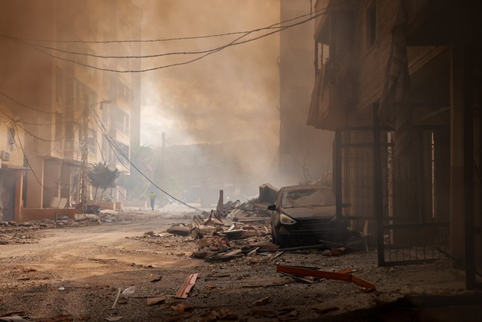 A pile of rubble and a dust-covered car in smoky street. One figure is walking along in the distance, wearing a hi-vis jacket