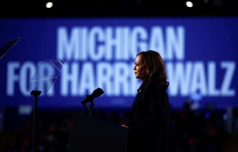 A woman with shoulder-length black hair wears a suit behind a podium. A large banner behind her reads "Michigan for Harris-Walz".