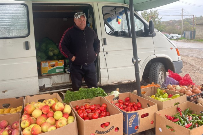 A fruit seller in Moldova stands next to a white van filled with produce. In front of him are boxes containing apples, tomatoes, and various vegetables. The van is parked on a gravel area with a small Moldovan flag visible inside the vehicle.