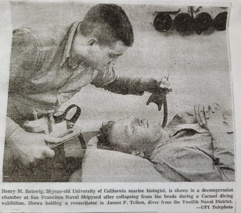 A black-and-white newspaper photo shows a man looking down with concern at another man who is lying with his eyes closed. 